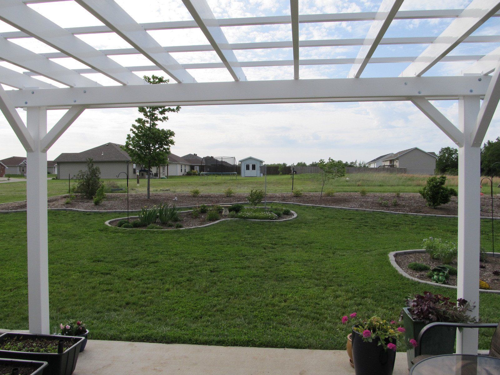 White pergola framing a view of a grassy backyard with garden beds and houses in the distance.