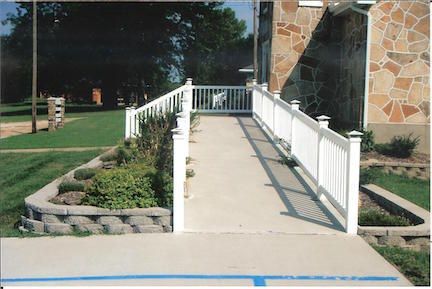 White ramp with railings leading to a building's entrance. Landscaping with greenery borders the ramp.