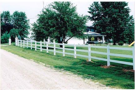 White fence bordering a grassy area next to a gravel driveway, with a house and trees in the background.