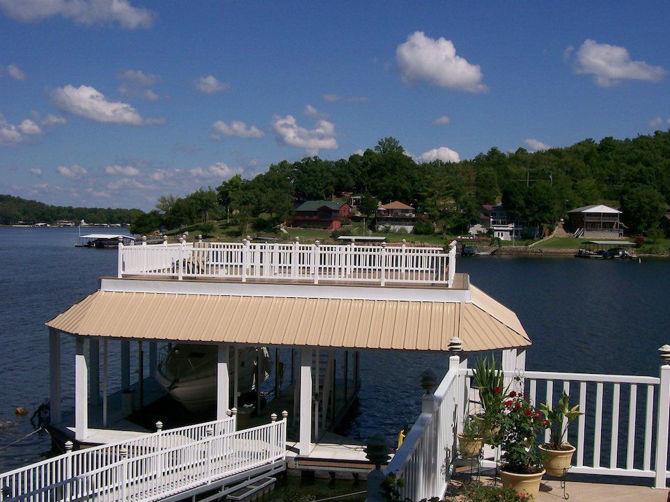 Dock on a lake with a white railing and a tan roof; trees and houses in the background under a blue sky.