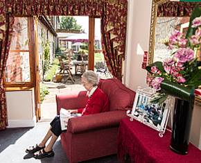an elderly women sitting on a red sofa and reading a paper