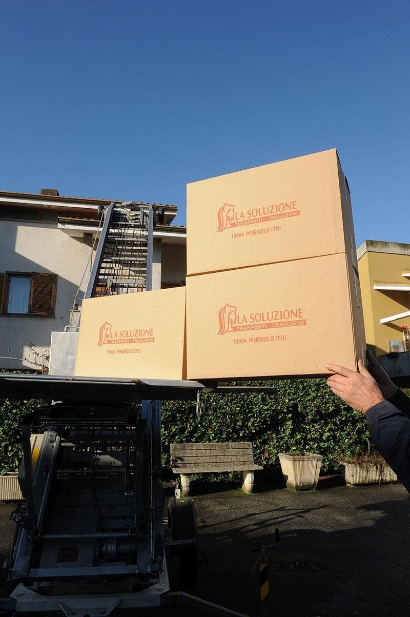 Scatole sollevate da un camion, impilate. Sullo sfondo un edificio e il cielo.