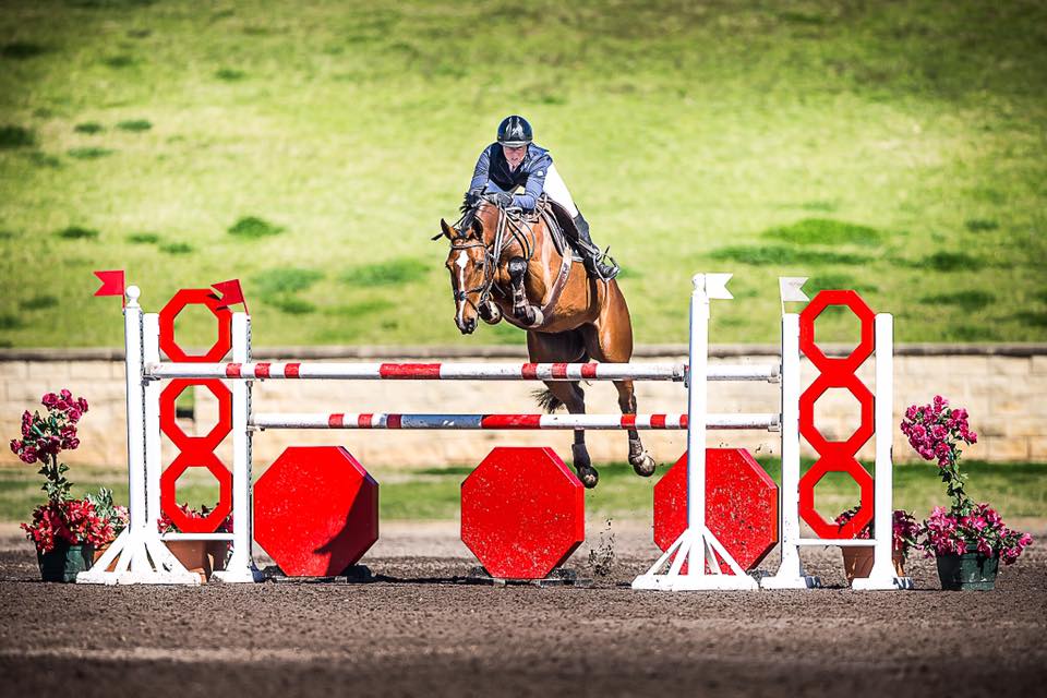 A woman is riding a horse over a red and white obstacle.