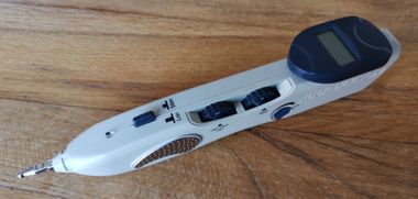 Een digitaal apparaat in penvorm met een klein lcd-scherm, diverse zwarte instelknoppen en een metalen punt op een houten oppervlak.