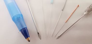 Een blauwe pen ligt naast diverse, slanke metalen medische naalden en instrumenten op een witte ondergrond.