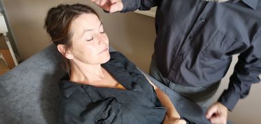 Een therapeut strekt een hand uit naar het gezicht van een persoon die met gesloten ogen ligt in een serene, binnenruimte.