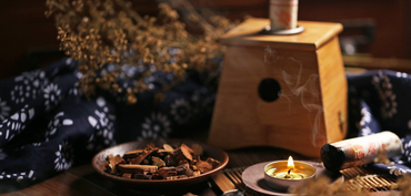 Een houten moxibustiebox produceert rook naast een kom met gedroogde kruiden, een brandende kaars en een donkerblauw doek met een patroon.