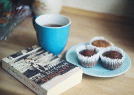 A cup of coffee is sitting on top of a book next to a plate of cupcakes.