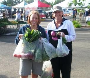 Two women standing next to each other holding bags of vegetables