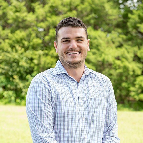 Man in a light blue plaid shirt smiles outside with green foliage in the background.