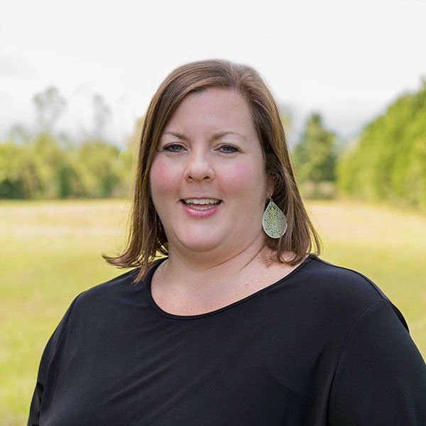 Woman with brown hair and smiling, wearing a black shirt and green earrings, in an outdoor field.