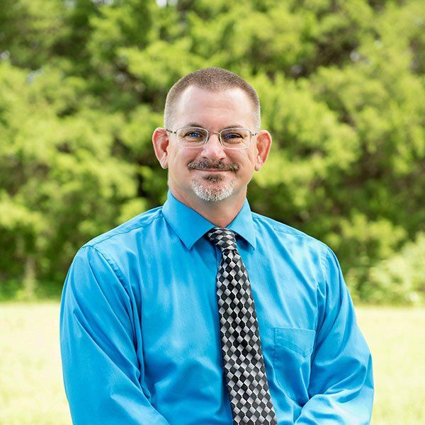 Man with glasses and a goatee, wearing a blue shirt and tie, smiling against a green background.