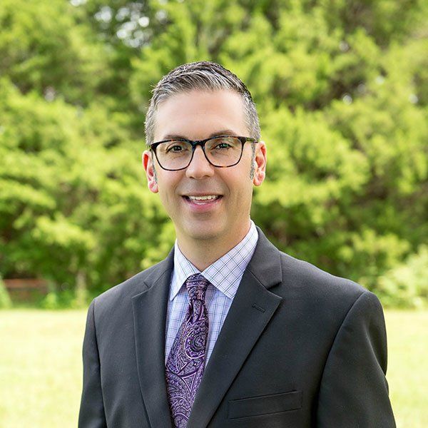 Man in suit and glasses smiling in front of green trees.