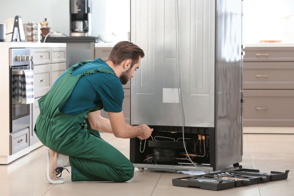 Réparateur travaillant à l'arrière d'un réfrigérateur dans une cuisine, vêtu d'une salopette verte.