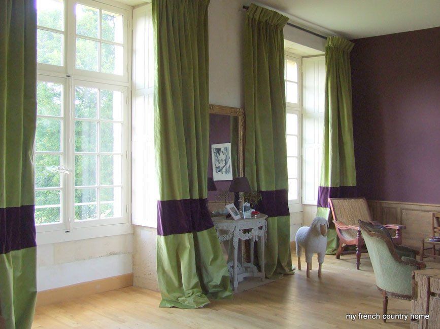 Stanza con grandi finestre, tende verdi con fasce viola e un piccolo cane bianco.