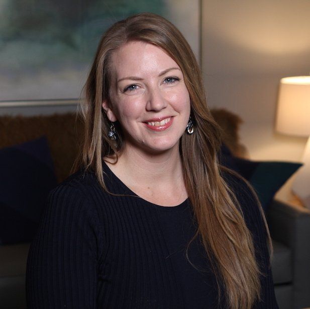 Smiling woman in a dark sweater sitting indoors near a lamp and couch