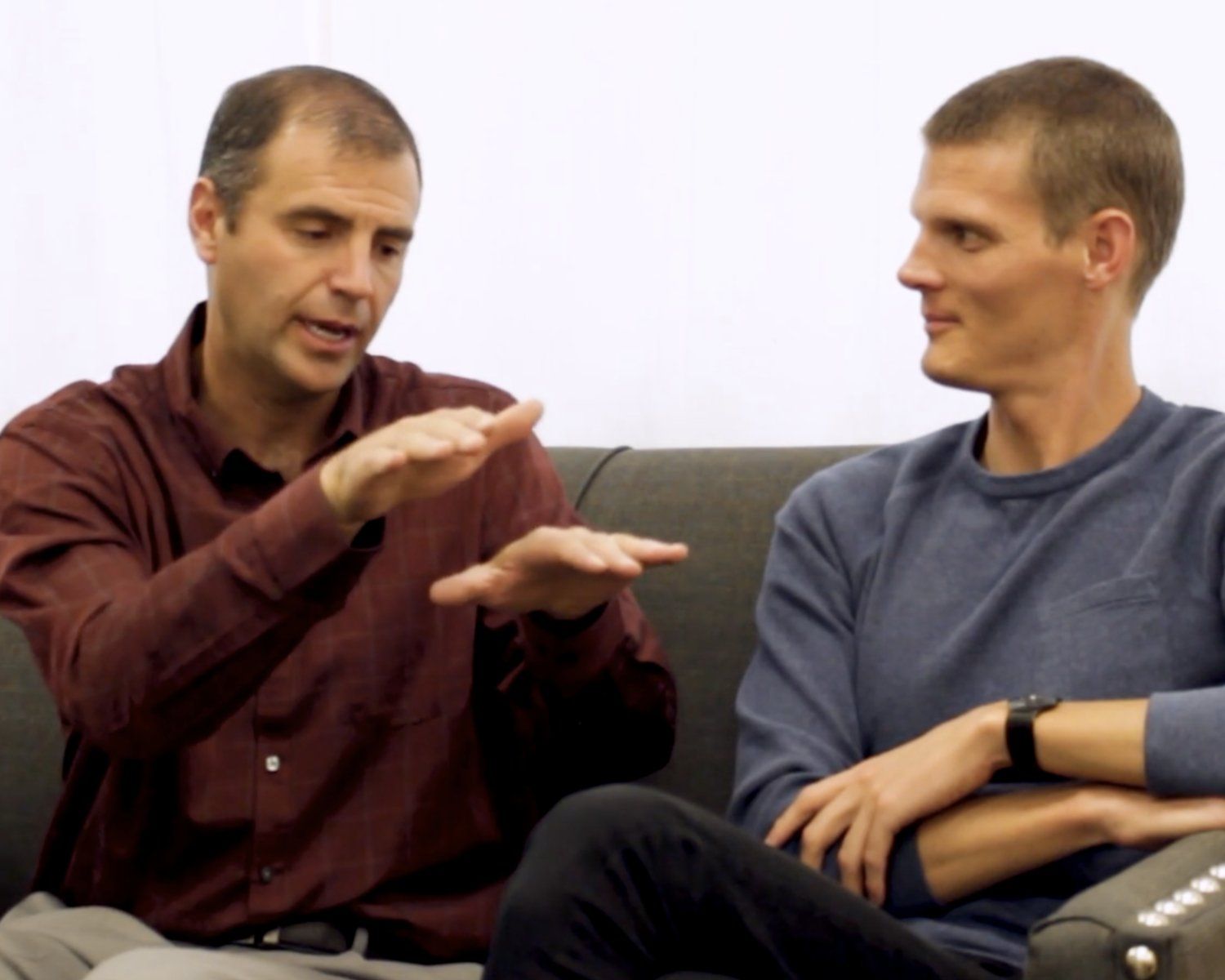 Two men seated on a couch, talking; one gestures with both hands while the other listens attentively.