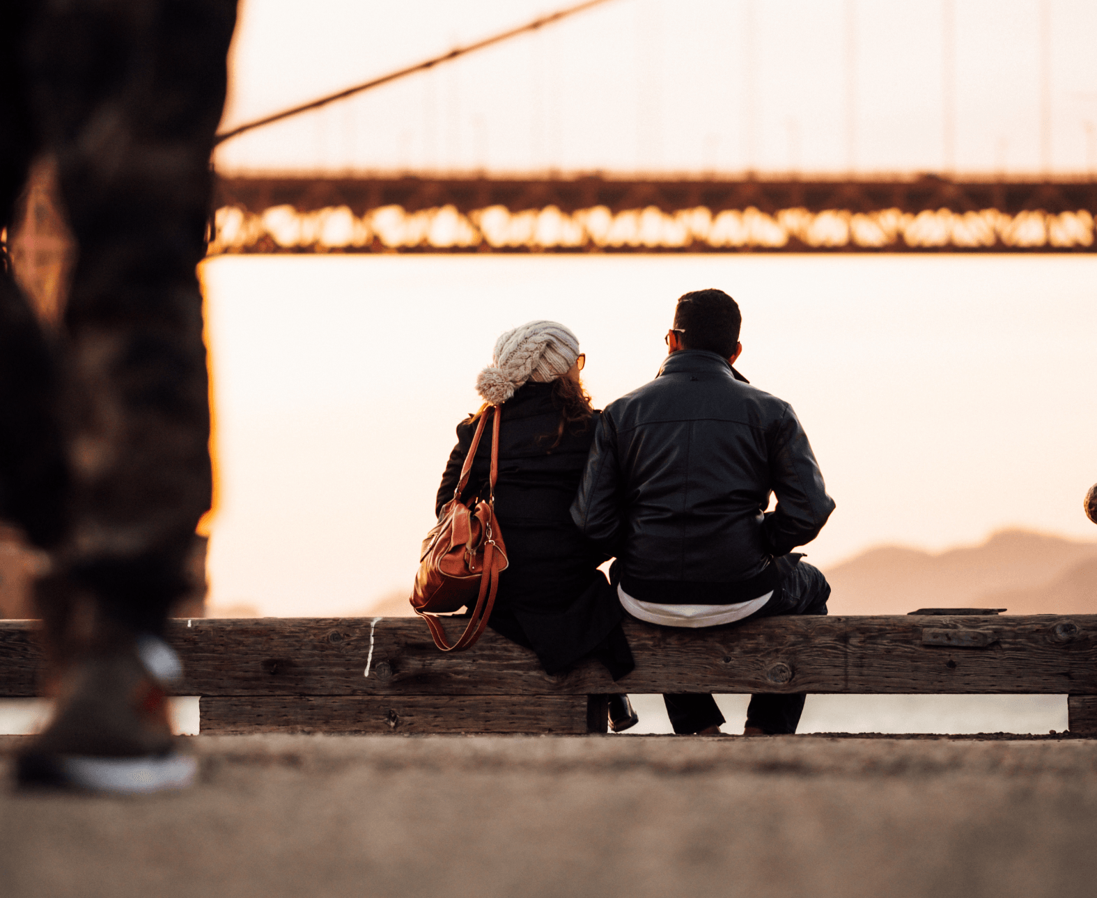 Two people sit on a bench by the water, facing a bridge at sunset.