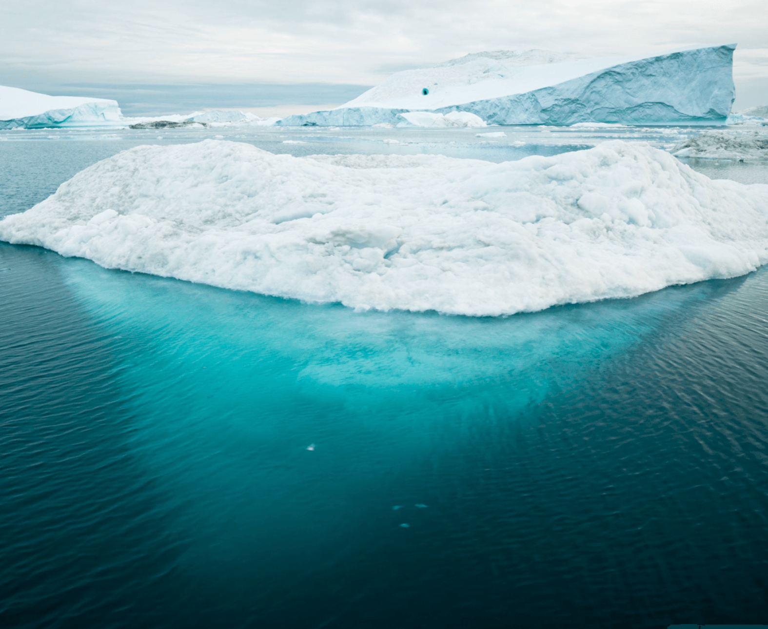 A large white iceberg floating in calm turquoise Arctic water under a pale sky