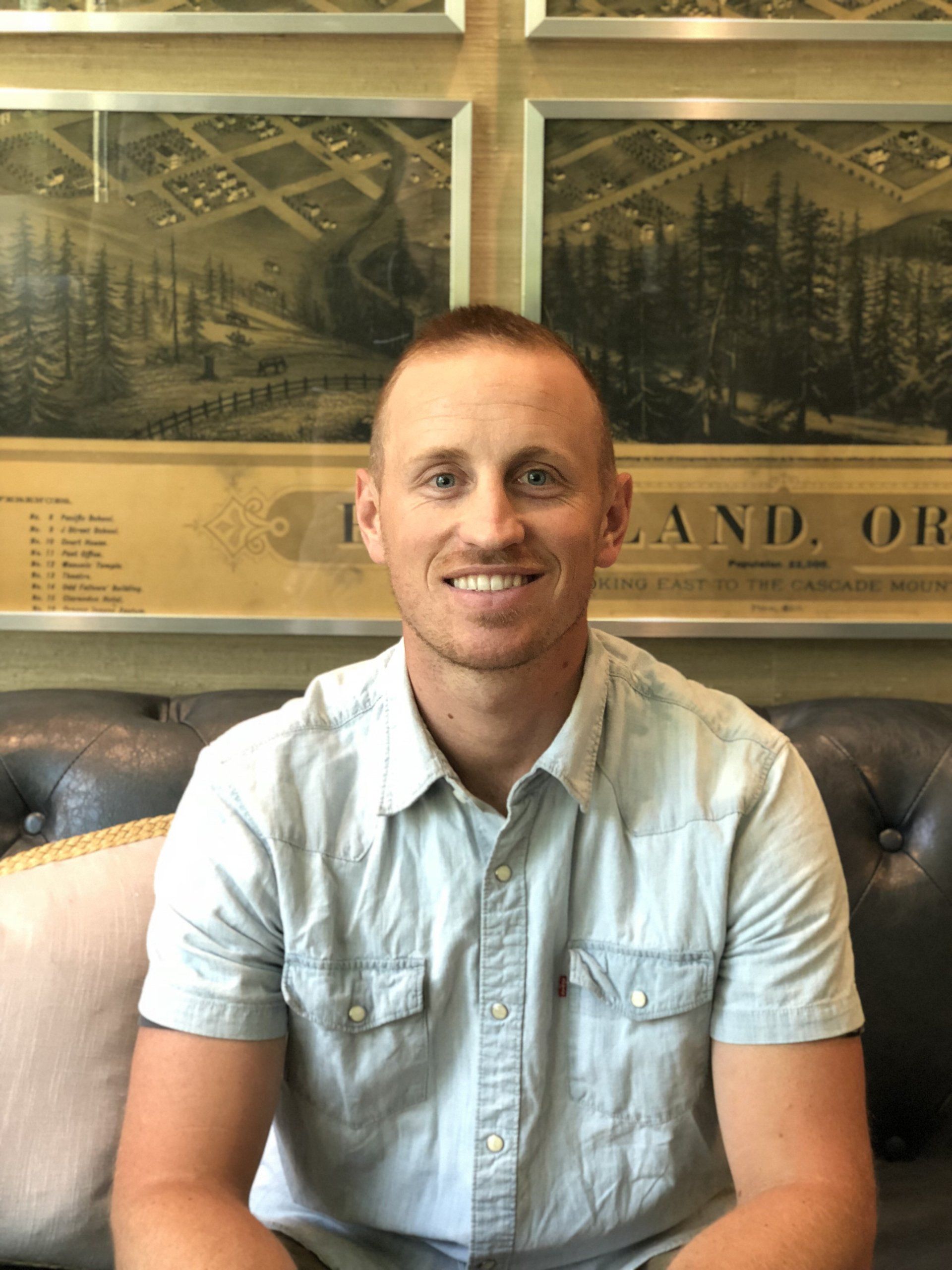Smiling man in a light blue shirt seated on a tufted couch in front of framed wall art.