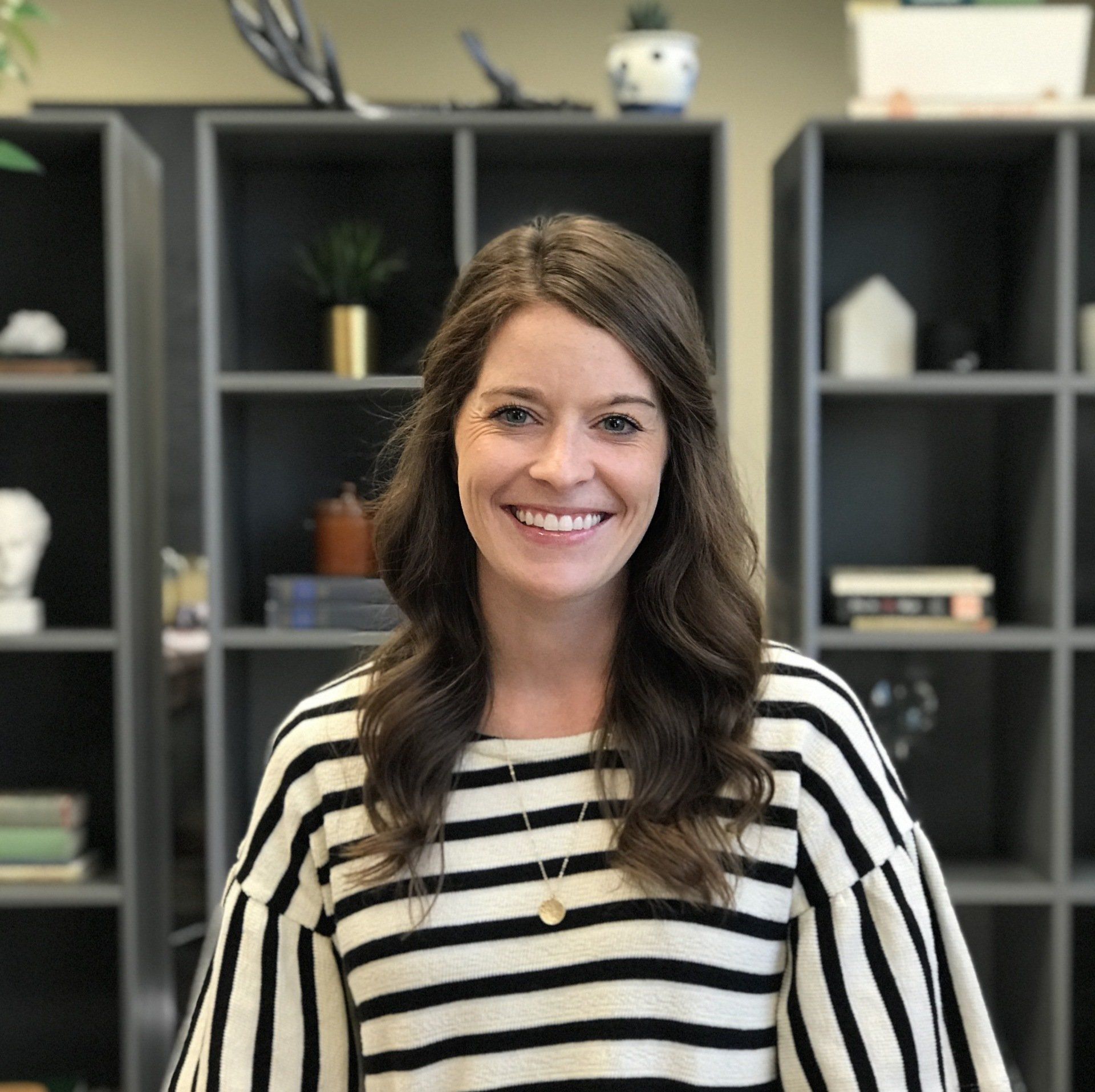 Smiling woman in a striped shirt standing in front of shelves in an office.