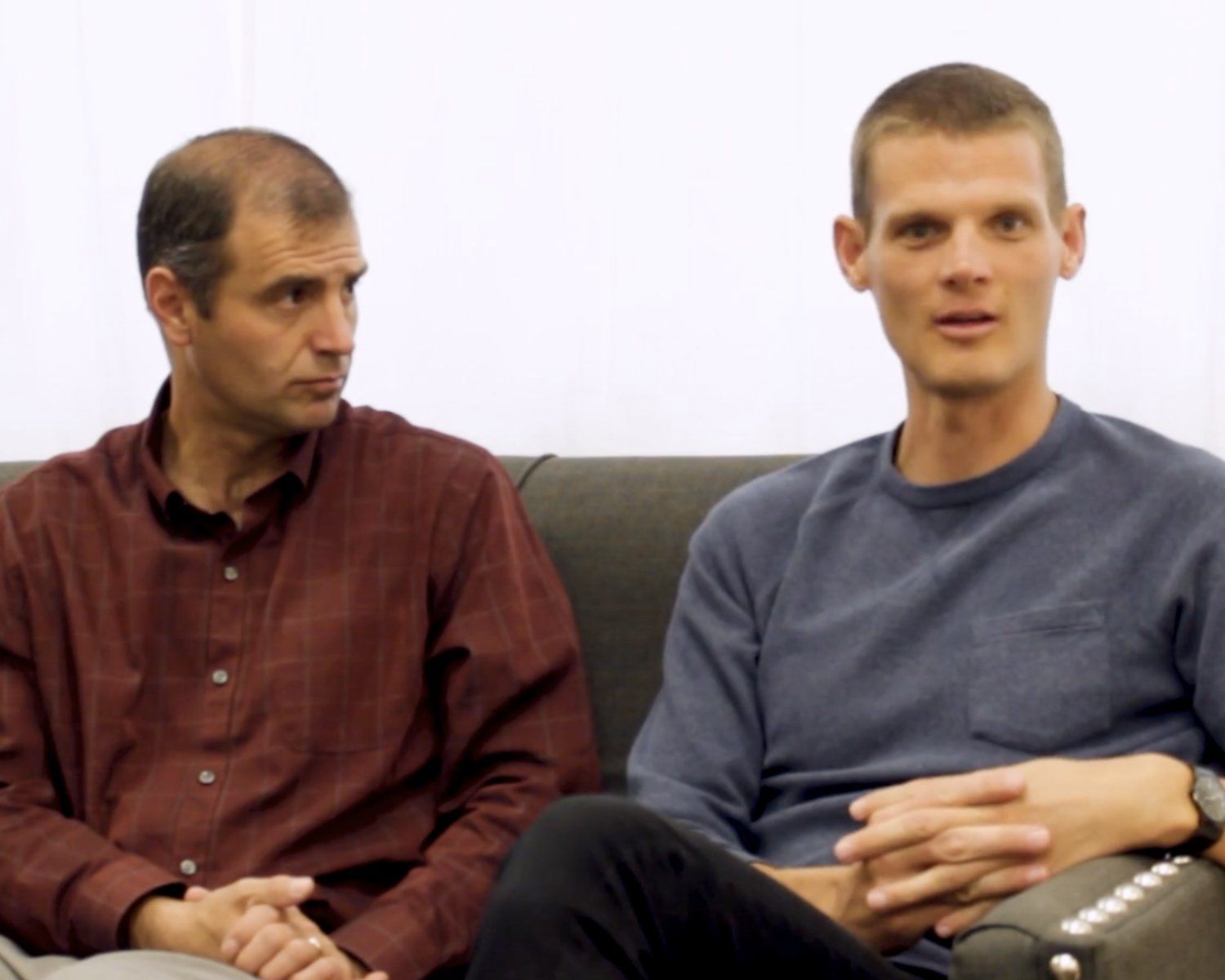 Two men seated on a gray couch, one in a red shirt and one in a blue sweater, in conversation.