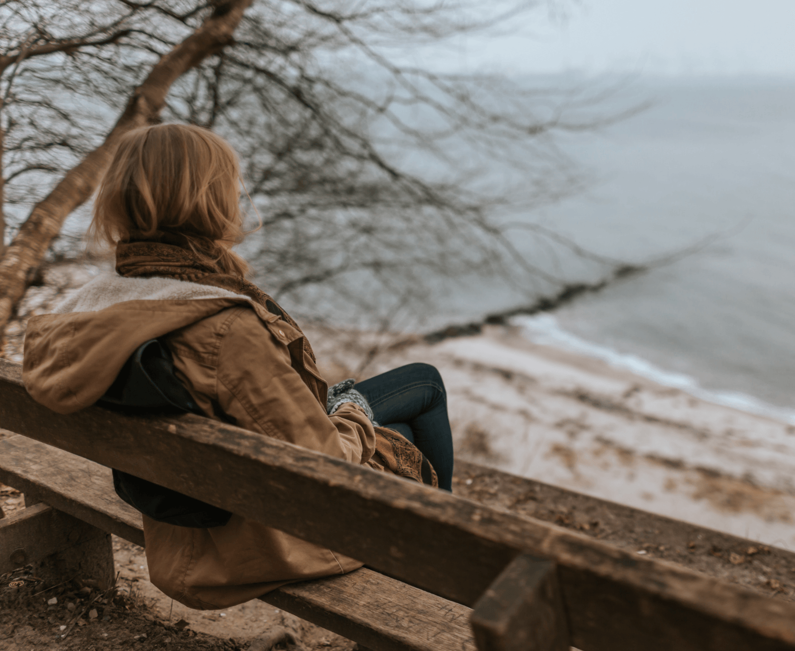 Person in a brown coat sitting on a wooden bench overlooking a cloudy beach.