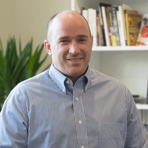 Smiling bald man in a blue striped shirt standing in an office with bookshelves and a plant