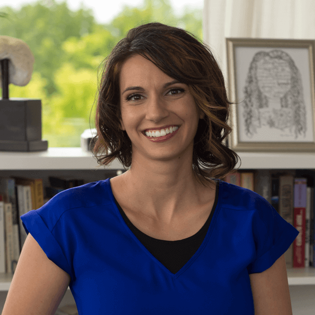Smiling woman in a blue top seated indoors with bookshelves and framed art behind her