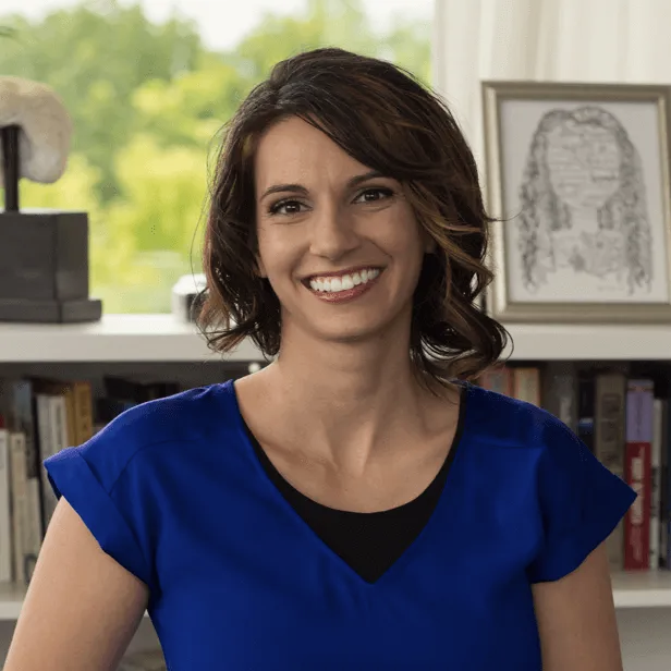 Smiling woman in a blue blouse in an office, with bookshelves and framed art behind her