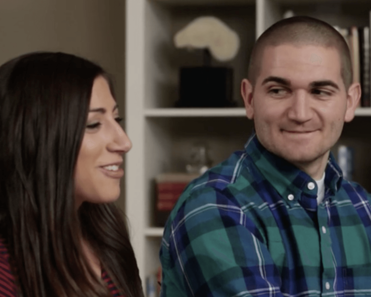 Two people smiling and talking indoors, with shelves blurred in the background.