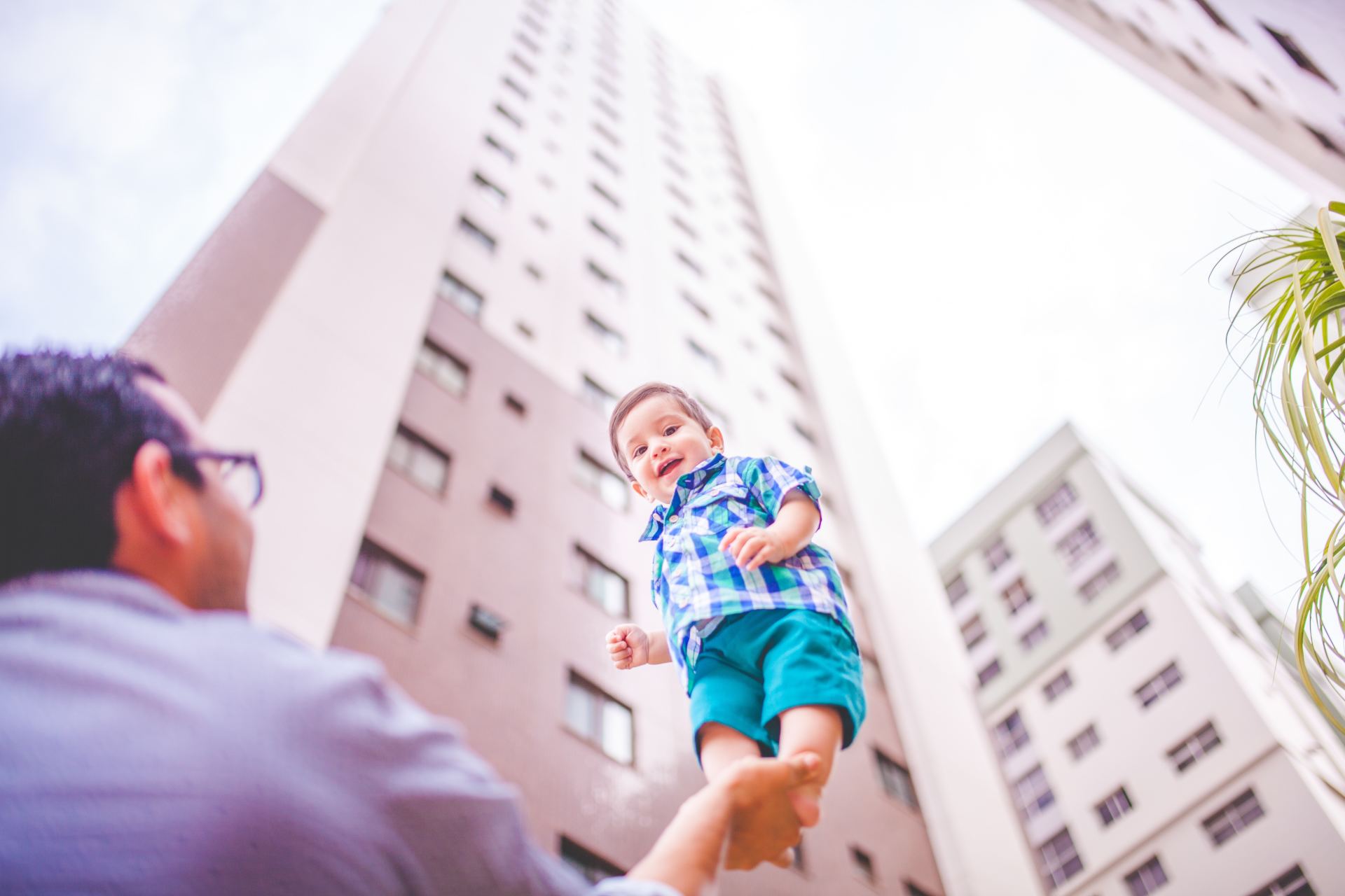 Adult lifting smiling child toward tall apartment buildings, low-angle view