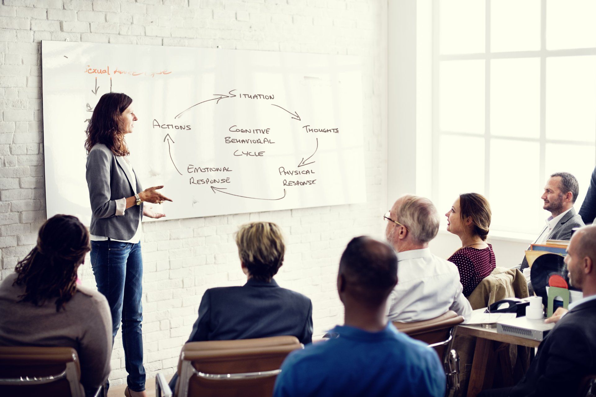 Workshop presenter speaking to a seated group in a bright classroom with whiteboard notes.
