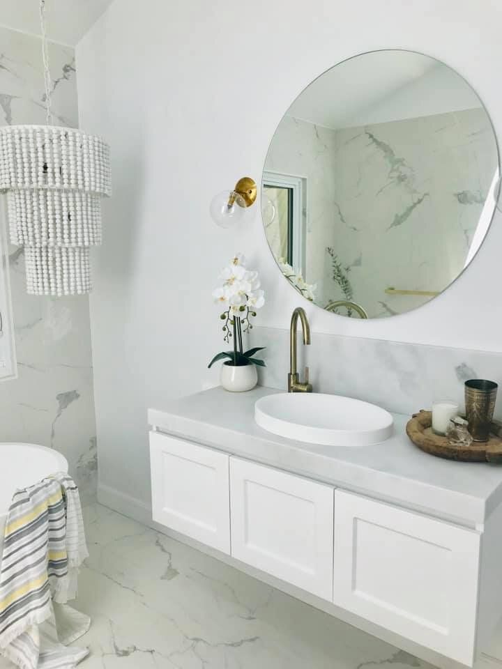 White bathroom with marble accents, floating vanity, round mirror, and beaded chandelier.