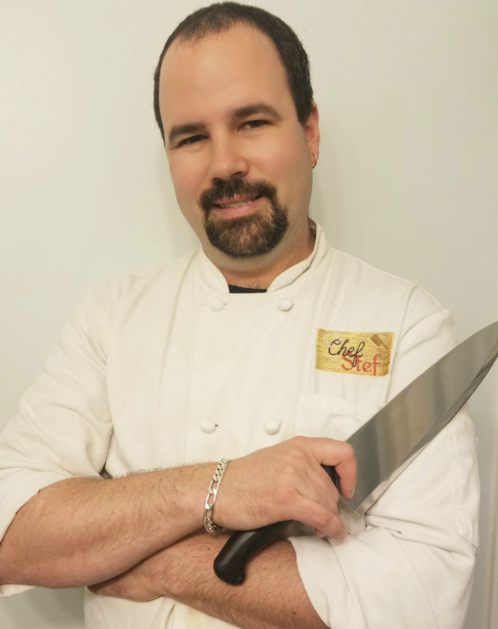 Chef in white jacket holding a large knife, standing against a plain background.