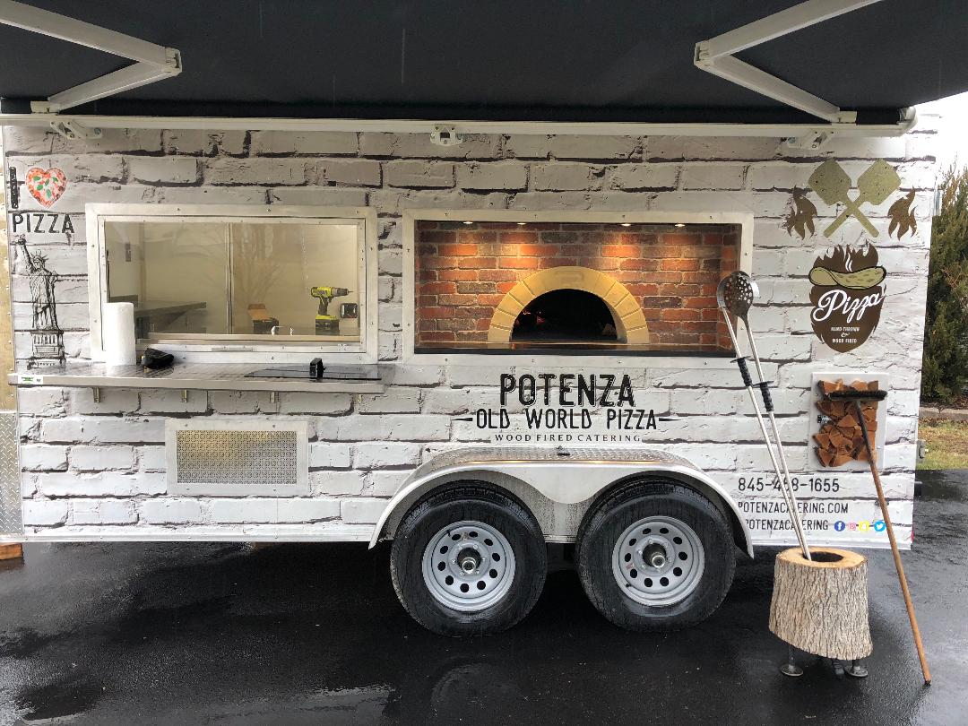 A food truck with a pizza oven on it is parked in a parking lot.