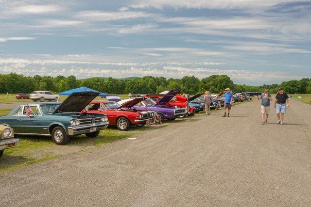 A row of cars are parked in a parking lot at a car show.