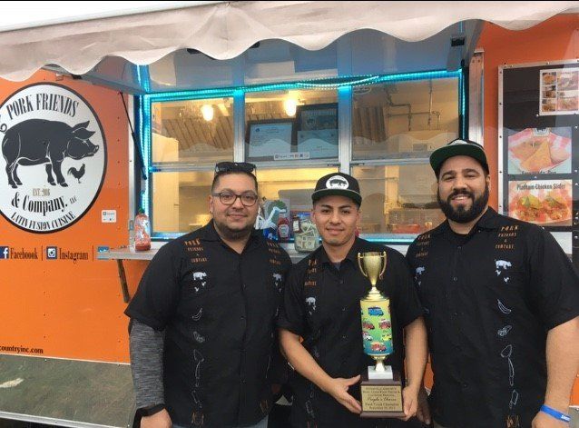 Three men are standing in front of a food truck holding a trophy.