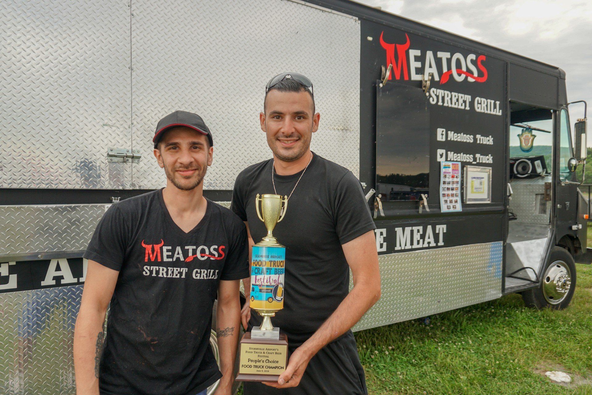 Two men are holding a trophy in front of a meat toss food truck.