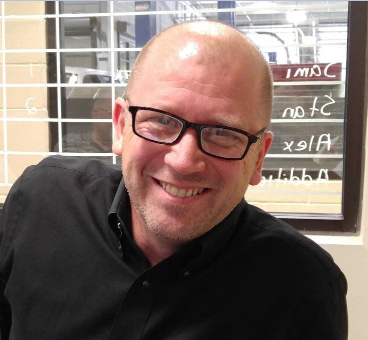 Bald man with glasses smiles widely, wearing a black shirt, in a room with a window behind him.