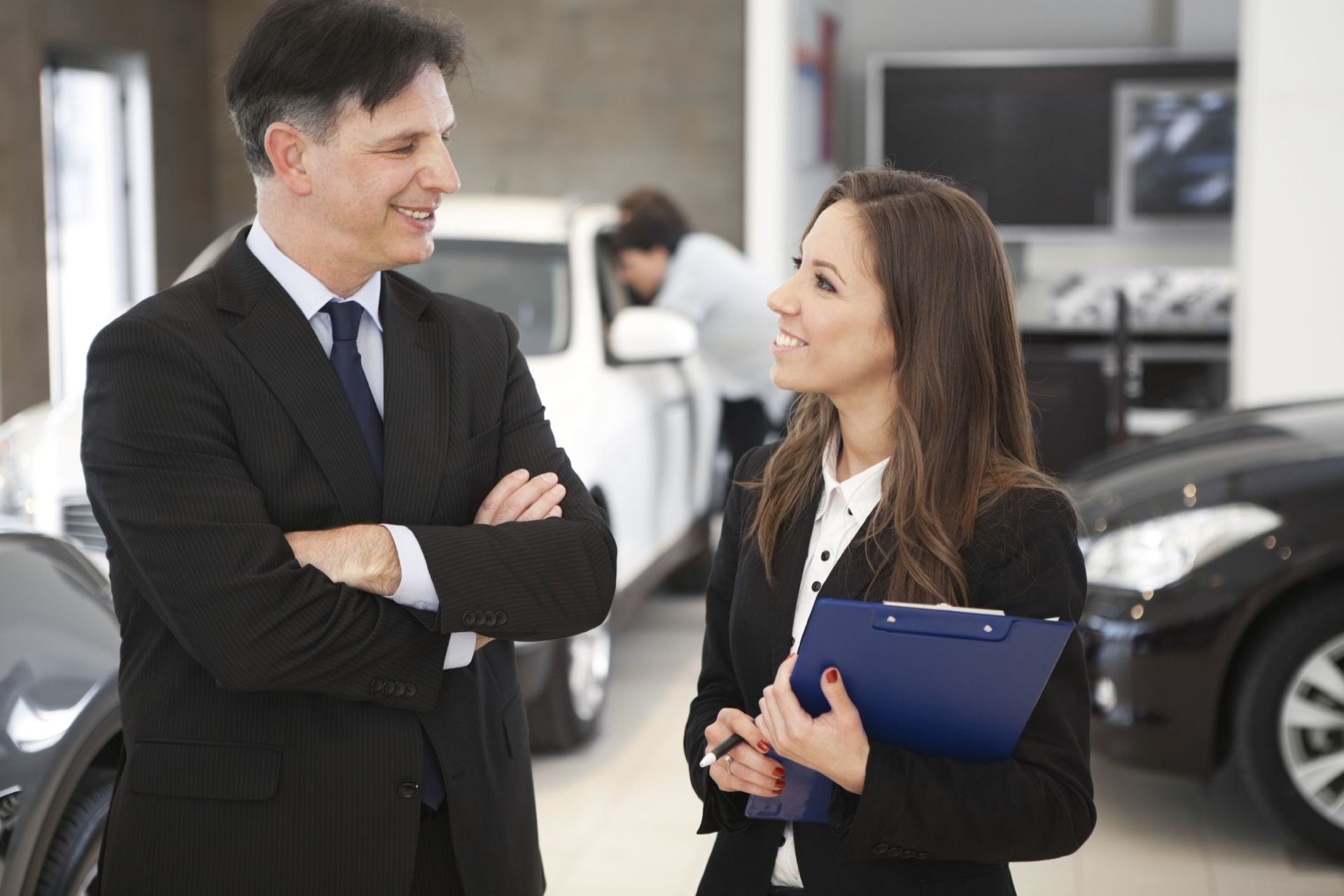 Car salespeople talking in a dealership, one with arms crossed, other holding a clipboard.