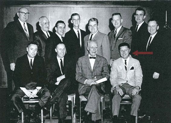 Group portrait of men in suits, some seated, some standing, in an office setting.