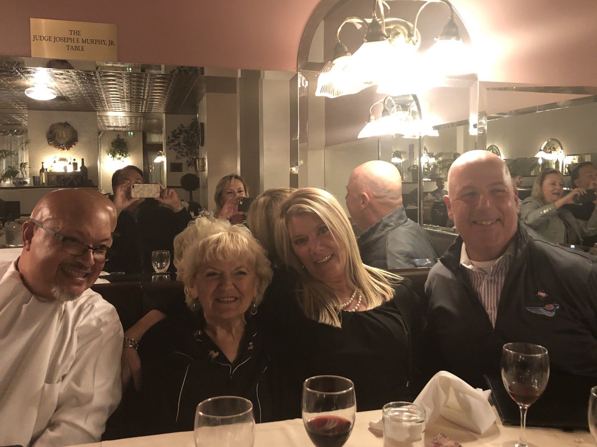 People at a restaurant table, smiling. Dark clothing, red wine glasses, and a mirrored wall.