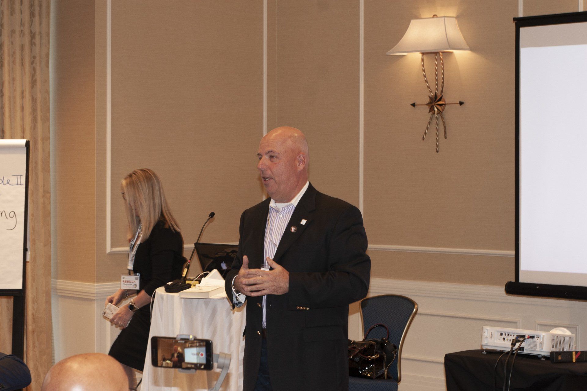Man in suit speaks at conference, woman stands nearby. Whiteboard and projector screen visible.