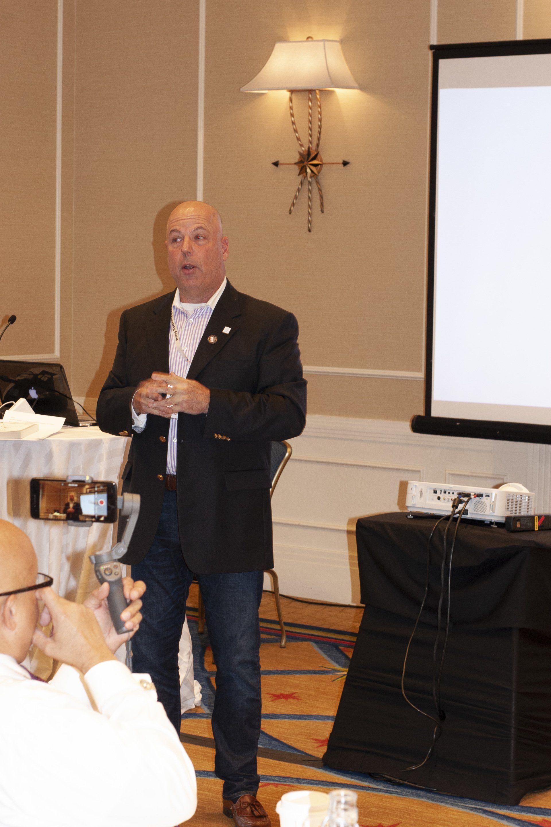 Man in a blazer gives a presentation. White screen behind him, with someone in the foreground taking a photo.