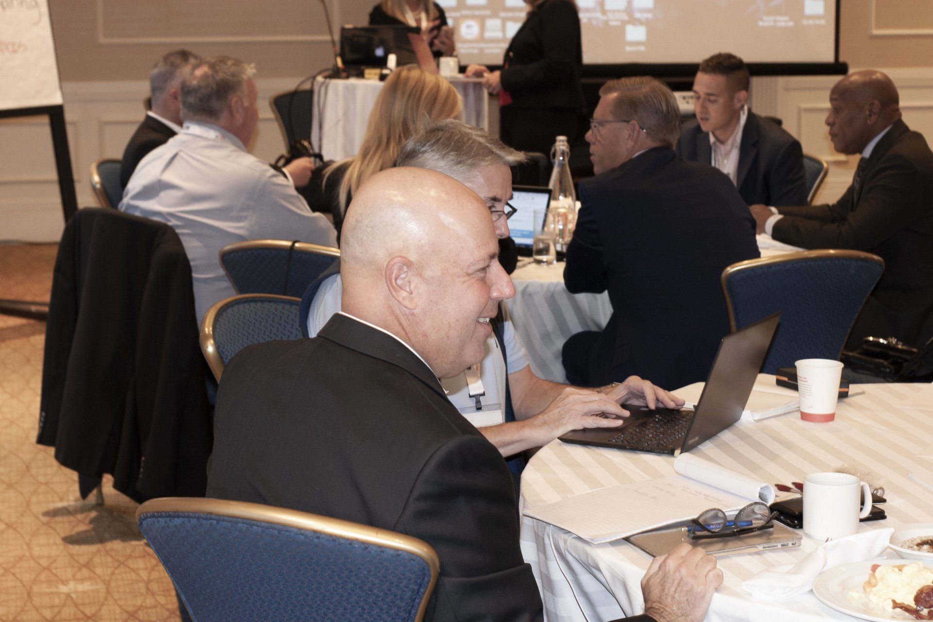 People at a conference, seated at round tables, some using laptops, engaging in discussions.