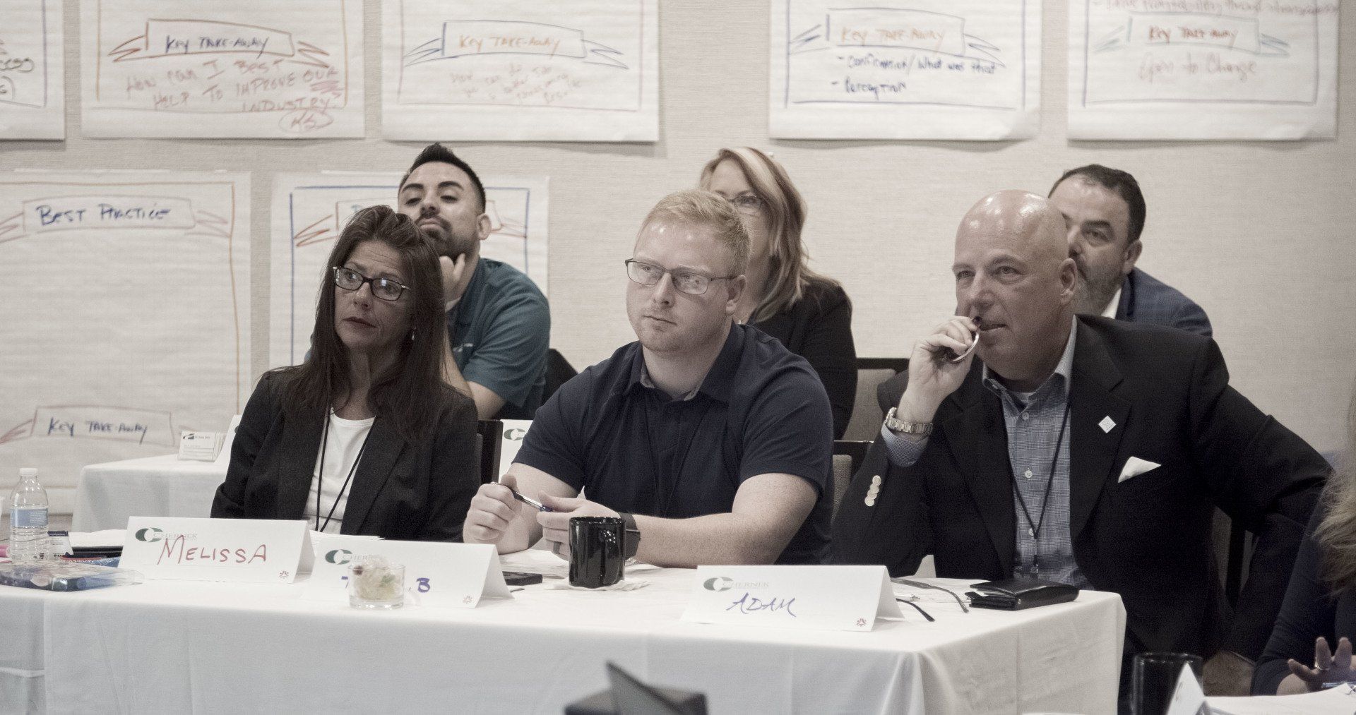 People seated at a table during a meeting; white papers with writing on the wall behind them.