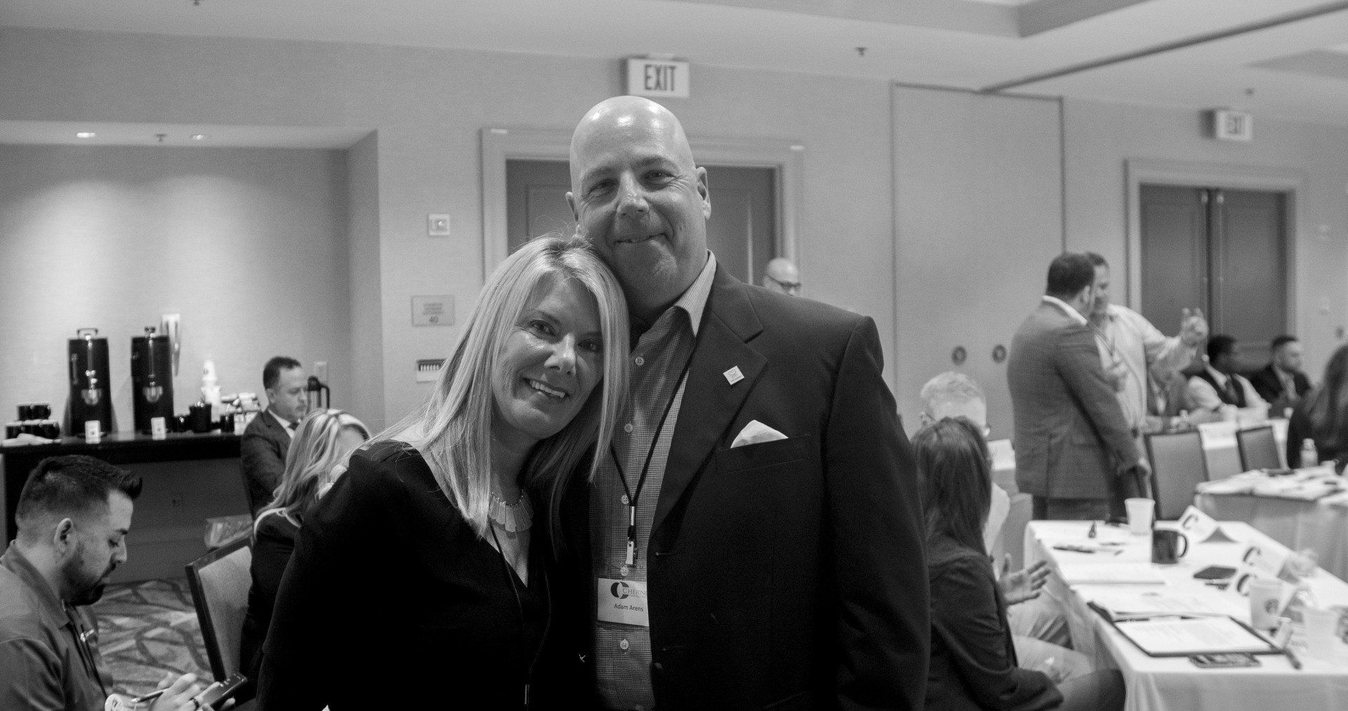 Man and woman pose together in conference room, smiling. Attendees in background at tables.