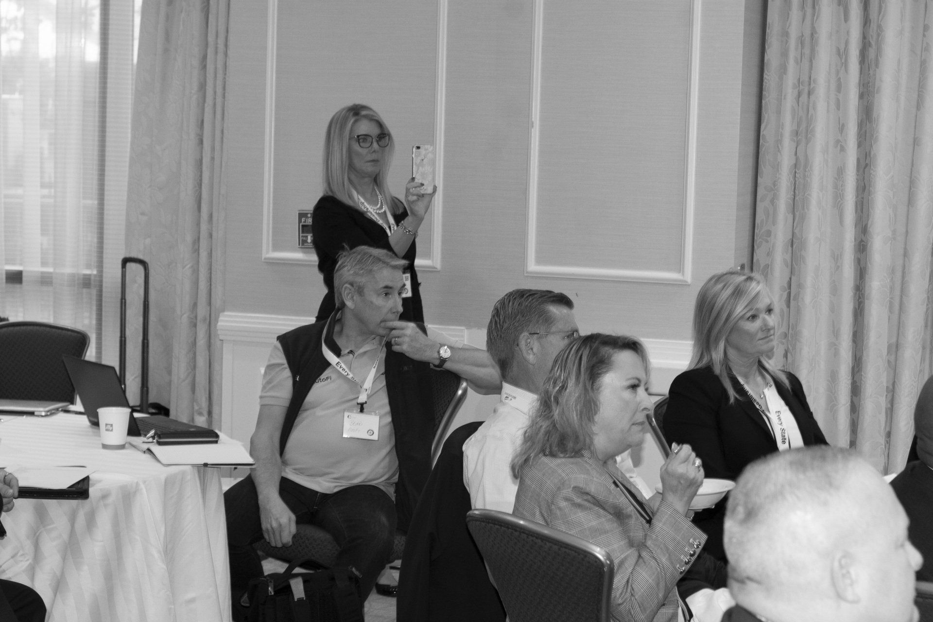 Attendees at a conference. One woman holds up a tablet while others listen. Indoors, near a table.
