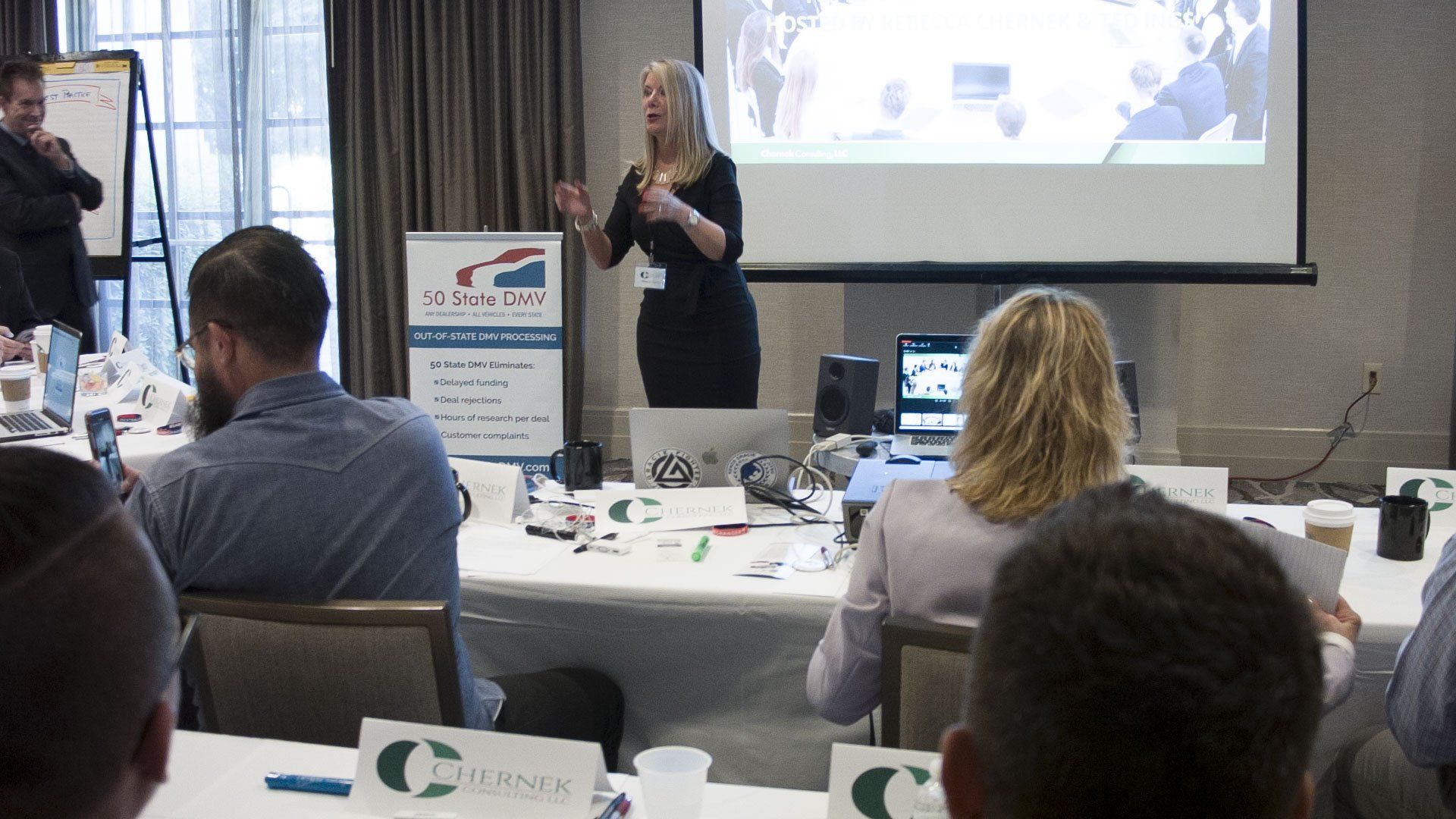 Woman giving a presentation in a meeting room, gesturing toward a screen; attendees seated at tables.